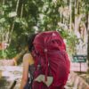 person with red trekker backpack standing on pathway surrounded with tall and green trees during daytime